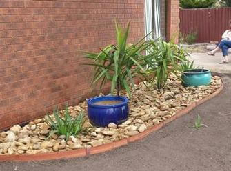 Completed garden bed Feature after preparation: Jarrah edge curved 75ml high fastened with steel pins, selection of builders plastic or weed mat to retard weeds, smooth large rivers peables, feature yakka's & clay pots.